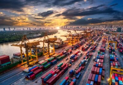 Container port with cranes and cargo ships at sunset, busy with shipping activity.